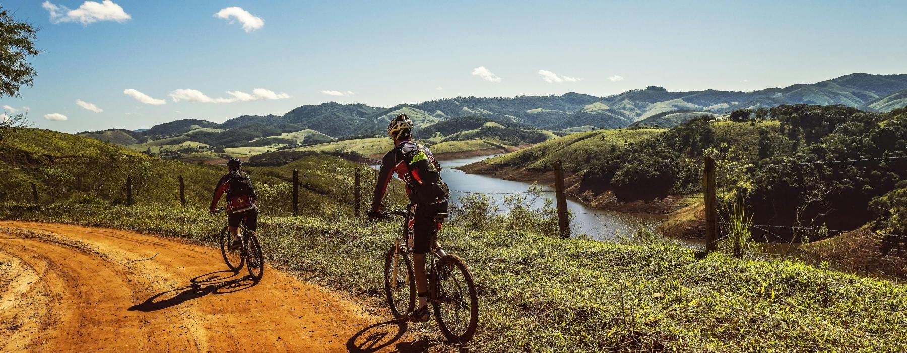 two people riding bikes on a dirt road near a river